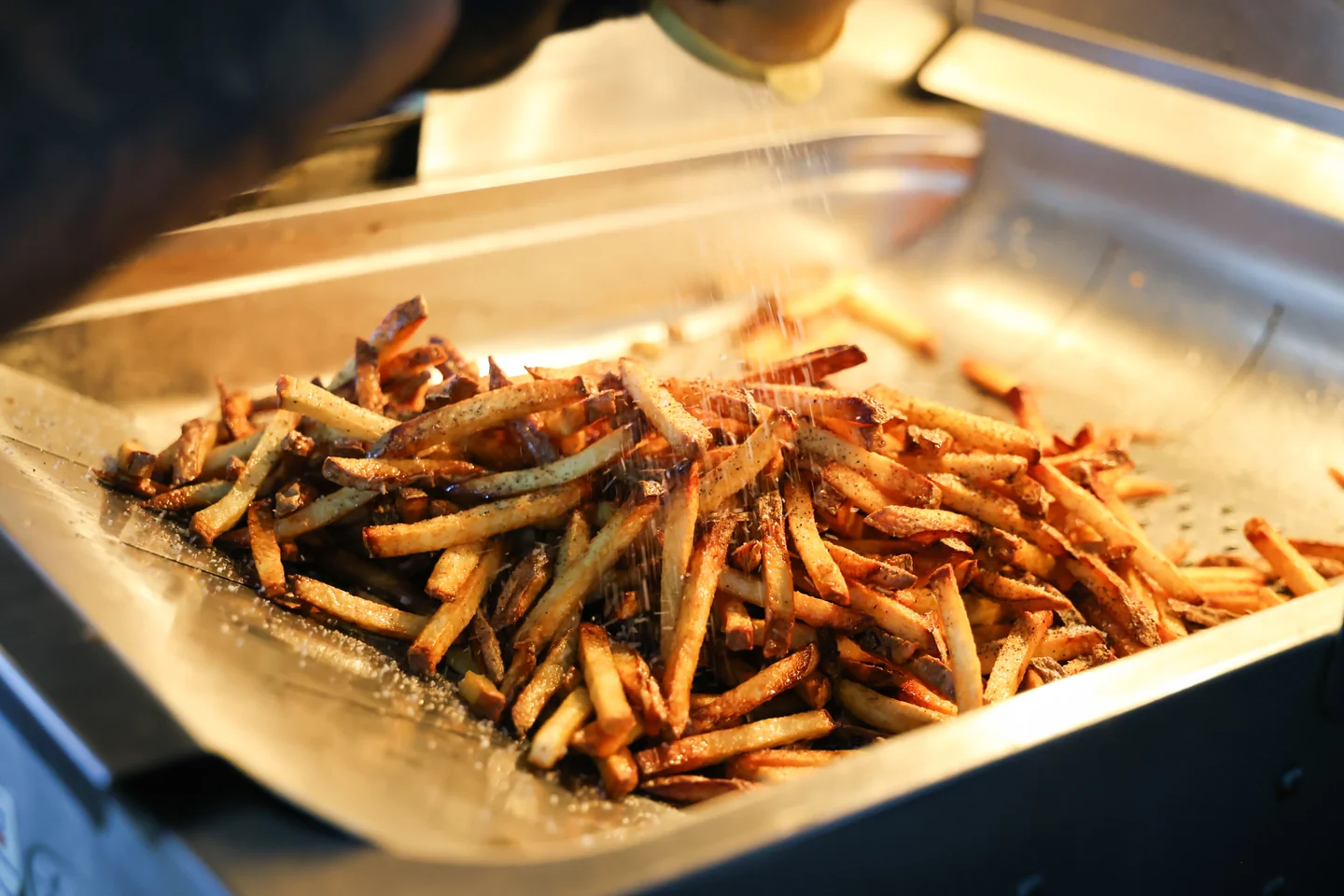 Fresh-cut fries being seasoned in a metal tray with steam rising